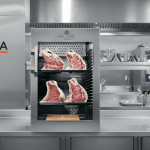 Armoire de maturation Dry Ager DX0500 Premium S posée sur un plan de travail, éclairage studio, grandes pièces de viande apparentes.