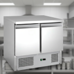 Saladette frigo inox 2 portes, équipement central d'une cuisine professionnelle, plan de travail en inox sous lumières de studio.